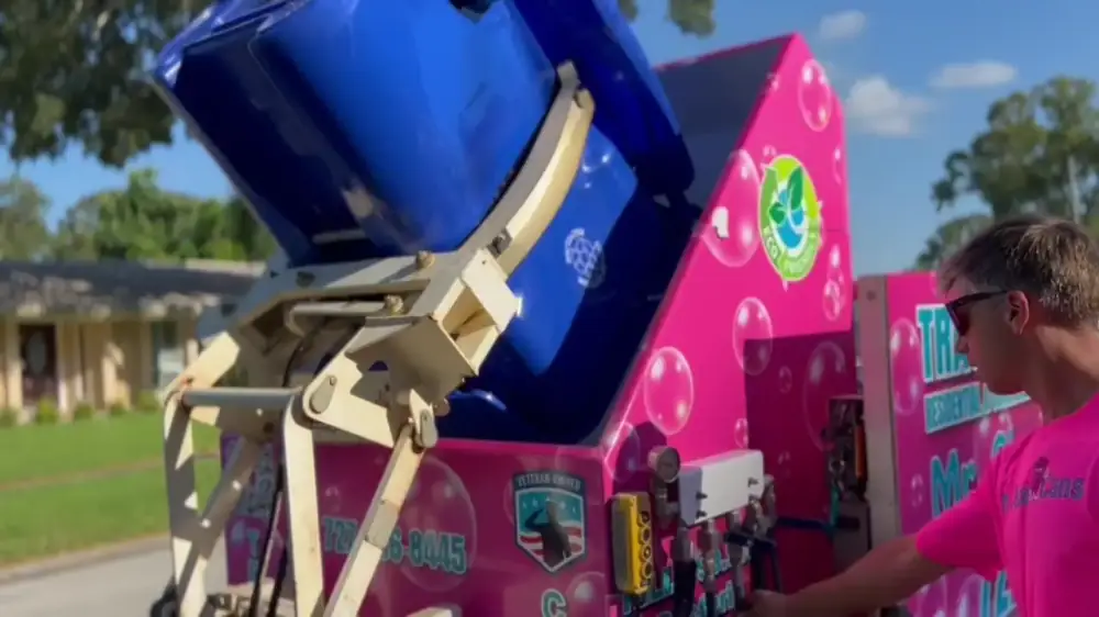 Mr. Clean Cans operator lifting a blue trash bin into the pink cleaning trailer with the bin-lifter arm